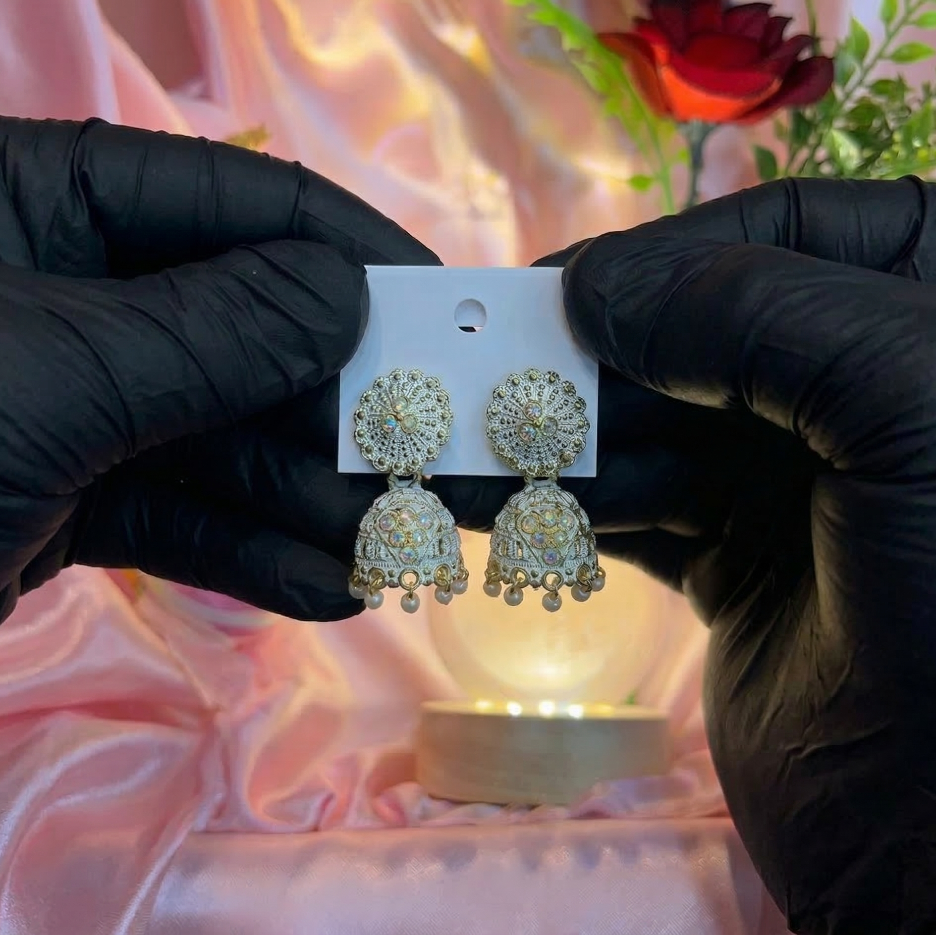 Gold earrings on a card held by hands in black gloves against a soft focus background with flowers.