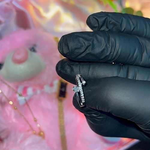 Hand wearing a black glove holding a diamond ring with a blurred background of a pink box and decorative items.